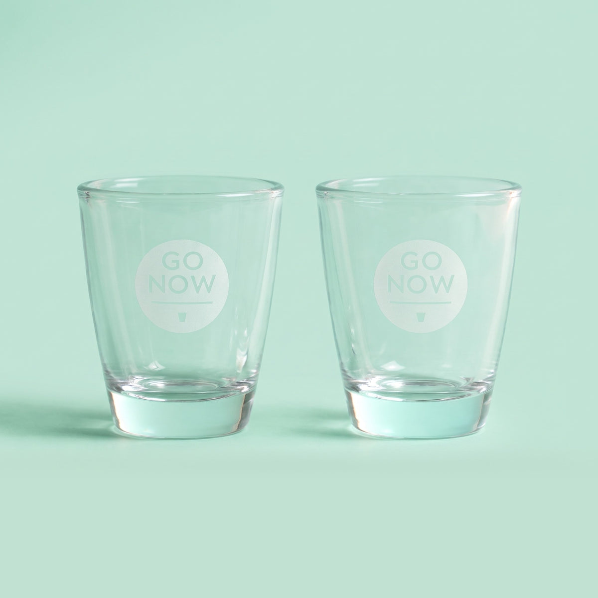 Twee lege Go Now shotglazen met gegraveerd logo op een mintgroene achtergrond