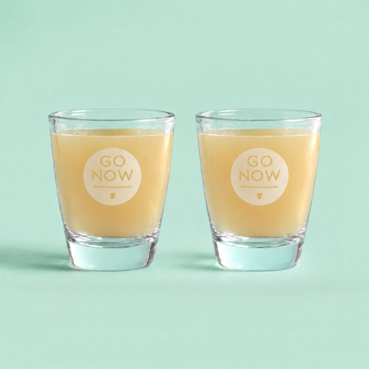 Twee Go Now shotglazen gevuld met gembersap op een mintgroene achtergrond