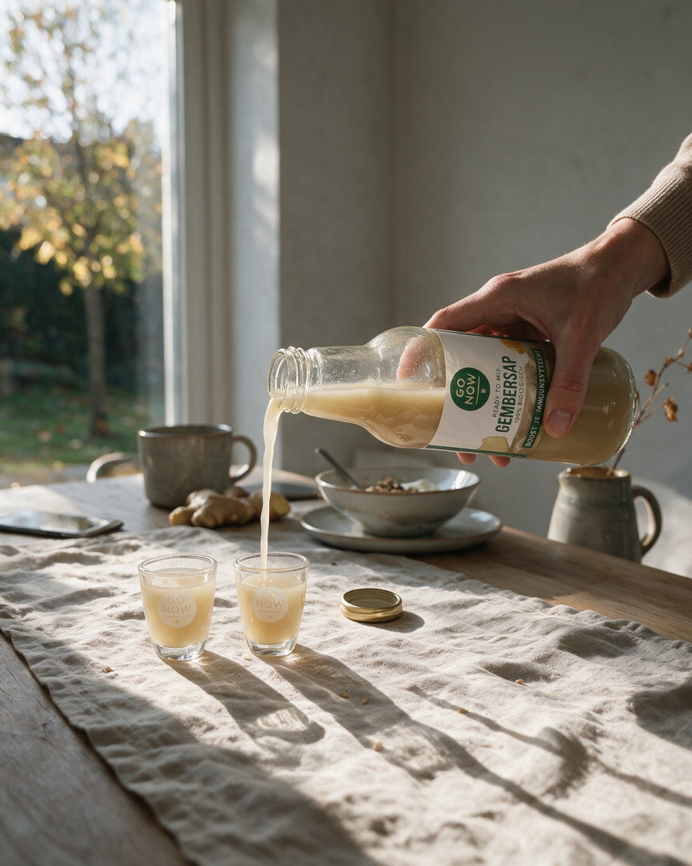 Hand schenkt Go Now gembersap in een Go Now shotglas op een linnen tafelkleed